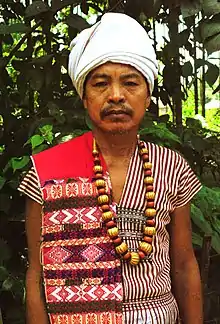 A Karbi man of West Karbi Anglong in traditional attire, wearing a Poho (white turban), a choi-hongthor (woven jacket), a lek paikom (gold-plated necklace) and another poho on his right shoulder.