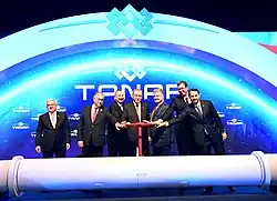Seven middle aged men in dark suits stand behind a large white horizontal pipe. There is a large TANAP logo behind them and six of them are holding the large red valve wheel protruding up from the pipe