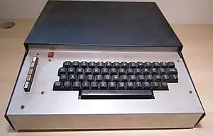An image of the Transam Triton home computer resting on a desk. The picture shows a keyboard in a long metal case with five function buttons to the left.