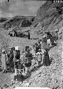 Line of men holding items of furniture in a hilly and rocky desert landscape