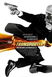 A white man in a black suit holds two guns on a white backdrop that has a band of orange. The words "The Transporter" is overlaid in the middle of the picture.