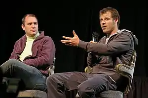 Two adult males sitting in chairs; the male to the right is speaking into a handheld microphone