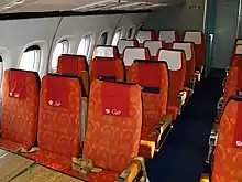 The passenger cabin of an airliner with orange seats and blue carpeting, the cabin is illuminated by daylight from the windows.
