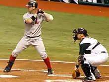 A man in a grey baseball uniform stands at home plate in a left-handed batting stance.