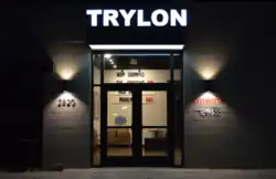 A dark storefront facade with the words "TRYLON" in illuminated white capital letters above the double glass doors. On either side of the doors, lamps cast triangles of light upward and downward to form the shape of an hourglass or bowtie rotated one quarter turn.