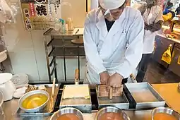 Man in chef's whites at a stove, cooking in four rectangular pans