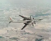Top perpendicular starboard view of four-engine propeller-driven aircraft in-flight. The pale gray/white aircraft is flying above almost white terrain.