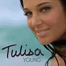 A close-up of Tulisa who is smiling in front of a blurry sea background.