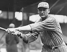 A man in a light-colored pinstriped baseball uniform looks into the camera having just swung his bat–the left.