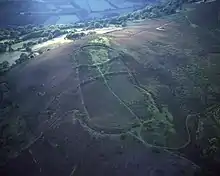 An aerial view of a mountain top showing an large oval ditch and wall split horizontally into three areas by two ditches and walls