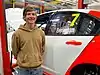 A teenaged man wearing a mustard hoodie standing next to a red and white racing car numbered 7 to the right of the picture