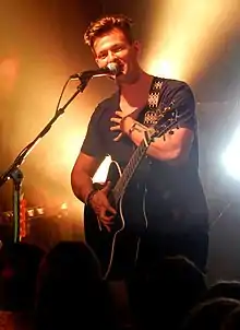 Tyler Ward wearing dark clothing, standing onstage with an acoustic guitar, appearing to sing or speak into microphone