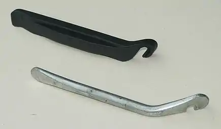 Two bicycle tire levers on a flat surface. The top one is made out of black plastic, the bottom one is made out of metal.