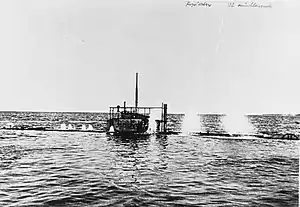 A photo taken from near the waterline of a submarine diving. Nearly the entire ship is underwater, with just the conning tower remaining above the surface. Spouts of water can be seen erupting on the surface from the submarine's air vents as it dives.