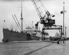 A photograph of a ship tied up to a pier. The bow of the ship points toward the left of the frame. On the right of the frame is an overhead crane.