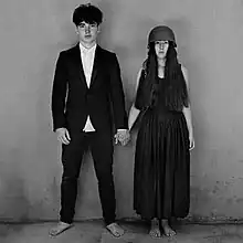 A monochrome image of barefoot young man in a dark suit coat holding hands with a barefoot young woman in a dark, ankle-length dress who is wearing a military helmet