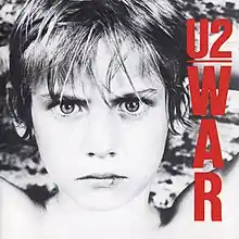 A black and white image of a young boy with his hands behind his head staring fiercely at the viewer. The bands name is seen in red text underlined while the album's title is placed on the right side and red vertically.