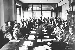 Photo of a caucus meeting showing thirty-five men and a woman, sitting around several long tables with documents laid out before them. As space is cramped and there is not room for everyone at the table, they are seated in two rows.