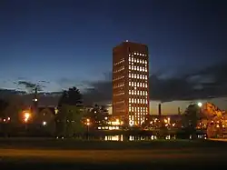 Photograph of a University campus just after sunset. A rectangular skyscraper dominates the view, standing in silhouette against a dark blue sky. To its left is a small, stone chapel with illuminated clock tower