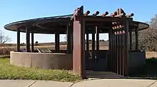 Circular roof on metal poles above circular concrete base; entrance made of metal poles set up like logs