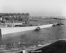 A black-and-white photograph of a battleship hull being moved across a harbor by tug boats.