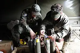 A group of 105mm artillery shells with plastic explosive stuffed into their fuze pockets to act as booster charges. Each of the 5 shells has been linked together with red detcord to make them detonate simultaneously. To turn this assembly into a booby trap, the final step would be to connect an M142 firing device to the detcord and hide everything under some form of cover e.g. newspapers or a bed-sheet.