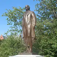 Bronze statue of a man in garb of the late 18th century. The statue is in an outdoor setting and the man is depicted as looking forward.