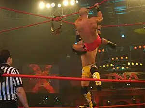 Two men, one in yellow trunks while the other in red trunks, battling, while hanging by red steel ropes, to retrieve a championship belt, which is suspended on the ropes