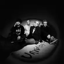 The image depicts the band sitting on the floor. The word "Unwanted" is written in dust on the ground.