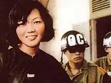 A Vietnamese woman dressed in black smiling on the left, next to two guards on the right wearing military uniforms and helmets