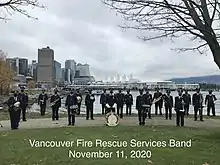 The band is pictured standing in two lines, each member spaced by about 6 feet. Each member is holding their instrument. Placed on the ground in front of the band and in the centre is the band's bass drum, which is decorated with the band name and the date 1927. Behind the band are grey, overcast skies, the water of Burrard Inlet, and, in the distance, the sail-like roofline of the Pan Pacific Hotel.