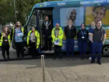 A blue and yellow 15 foot bus with several healthcare staff wearing blue scrubs and support staff wearing high disability coats. The local mayor wearing his chain of office.