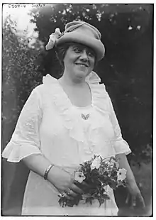 A woman standing outdoors, smiling, wearing a hat with a turned-up brim and a white dress with a ruffled collar and cuffs. She is holding a small bouquet of flowers.
