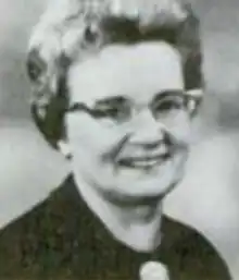 A smiling white woman with a bouffant blond hairstyle, wearing glasses and a dark top
