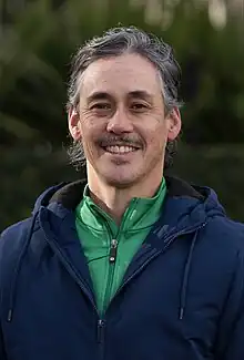 A middle-aged man with mid-length greying hair and who is wearing a green and blue jacket smiles at the camera