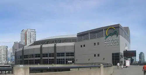 A grey building with two distinctive sections. The section on the left curves in a circular manner away from the viewer. The lower half is composed of windows. The section on the right is shaped like a square and has windows only in the bottom-left corner. A yellow circle and two jagged green shapes resembling triangles are at the top-right of the square building, with the words General Motors Place written in white capitals directly beneath. Several skyscrapers are visible in the background.