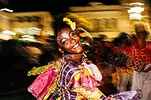 In the foreground, a black woman in bright-coloured carnival clothes smiling in the midst of dancing; a blurred background showing a crowd of people, lit-up buildings against a night sky, and a few other women of color in carnival clothes to the right