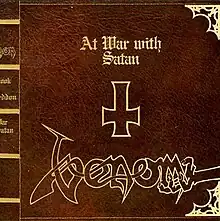 Cover resembles a leather bound book. All design elements are in golden print on top of the leather background. The design includes a pseudo-spine and pretend metallic corner protectors. In the center of the cover is an inverted cross with the title "At War with Satan" above in a gothic-like font with the "Venom" band logo below stylized to include a clawed demonic hand reaching in to grab the "m" from the right.