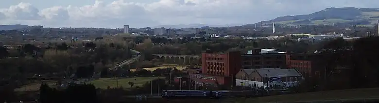 View towards Glenrothes seen in its landscape setting from a nearby cemetery. A train is leaving nearby Markinch Station on the East Coast Mainline. Glenrothes town centre with the numerous taller residential and office buildings can be seen in the centre of the image. The River Leven Bridge provides a stark white vertical emphasis on the right side of the image. The Lomond Hills regional park and rolling countryside form the backdrop on the horizon