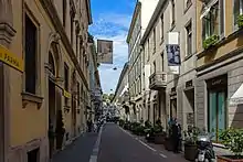 A narrow street with multi-story buildings in mostly different shades of yellow, with blue sky and clouds above. The building along the left near the camera has signs with the Acqui di Parma logo, and Cartier is along the other side. There are some people walking along the sides of the street in the distance