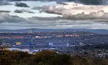 Trees in the foreground; houses, shops and industrial buildings in the middle ground; and hills in the background. The sky is grey.