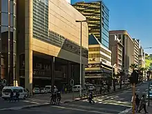 A busy, divided urban street seen from its right side, looking uphill to blue sky and a green hill in the distance. Tall buildings line the far side; the one nearest the viewer is too high to be contained within the image. Near the viewer is an intersection with a traffic light, with pedestrians crossing and waiting to cross