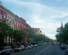 A street with three-story brick rowhouses in a variety of colors and short trees on the left, with taller trees and a distant steeple on the right.