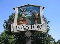 Closeup of village sighn, with a blue sky and green tree in the background.  The sign is framed like a medieval barn, around a low relief carving, painted in realistic colours, featuring a stream with a Roman soldier on the right and an 18th century smock mill on the left.