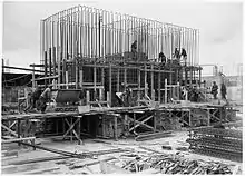 Scaffolding surrounds a half-finished concrete foundation. Dozens of metal steel poles rise from the foundation. A dozen workmen are visible and involved in various construction tasks.