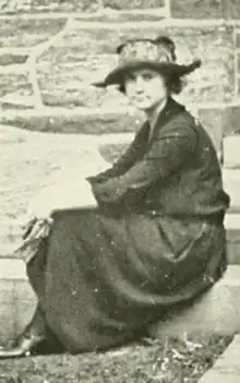 A young white woman, seated on a step outdoors, wearing a dark blouse and long skirt, and a hat with a floppy brim