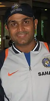 Virender Sehwag wearing a grey jacket and a cap, smiling.