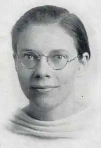 A young white woman with short dark hair parted on the side and combed away from face, wearing round eyeglasses and a light-colored top with a draped neckline