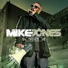 The cover shows a man wearing a black jacket, grey t-shirt, blue jeans and sunglasses in front of an apartment building. He's holding and speaking into a megaphone with light shining behind it.
