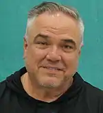 A 47-year-old man with short, gray hair, smiling at the camera with his head tilted.